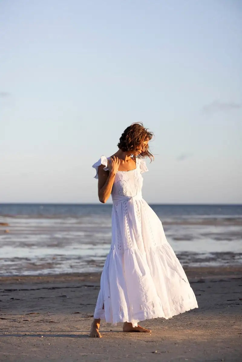 A flowing white embroidered maxi dress with ruffled off-the-shoulder sleeves and tiered skirt.
