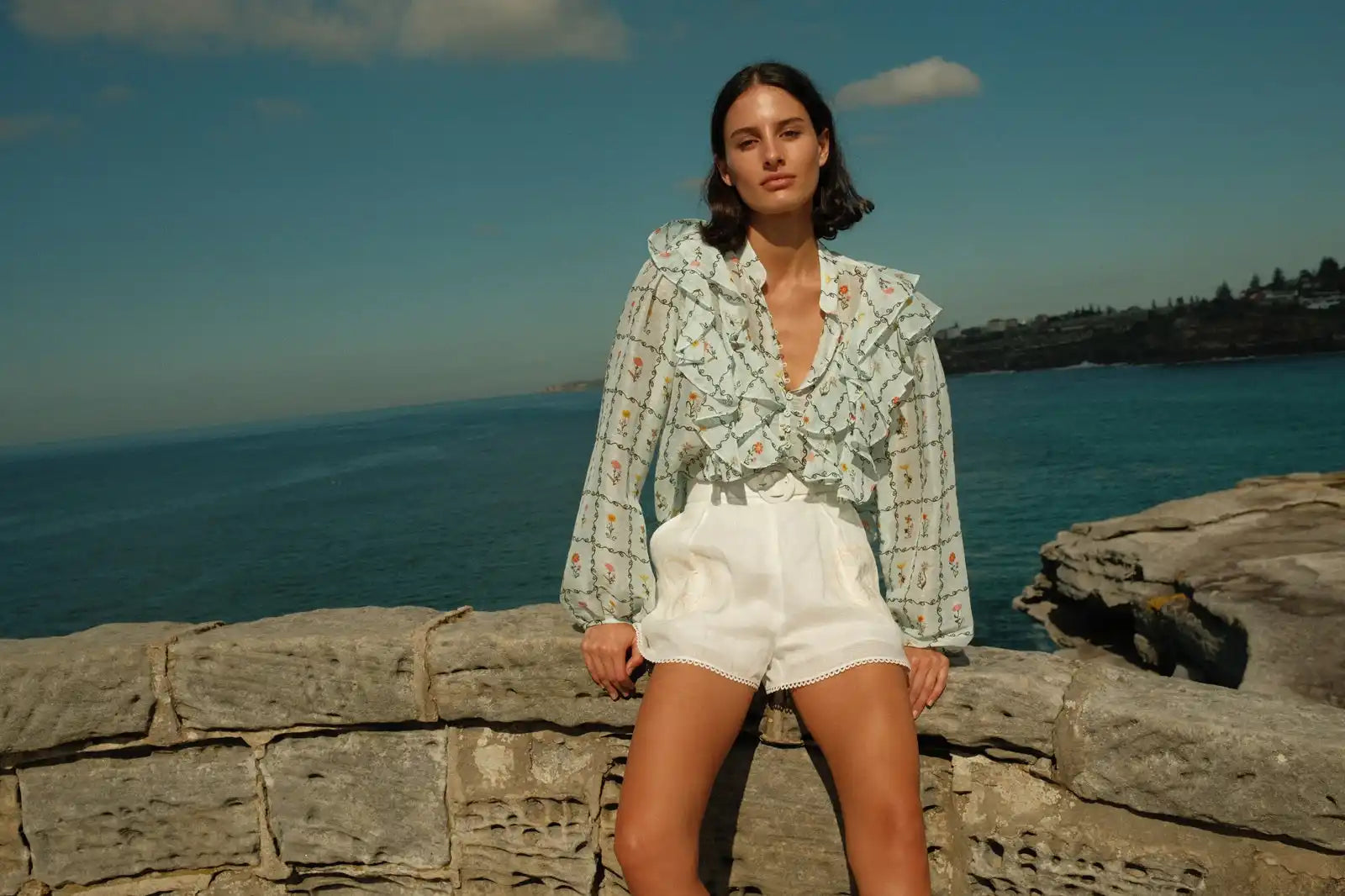 A woman wearing a light blue patterned ruffled blouse and white shorts.
