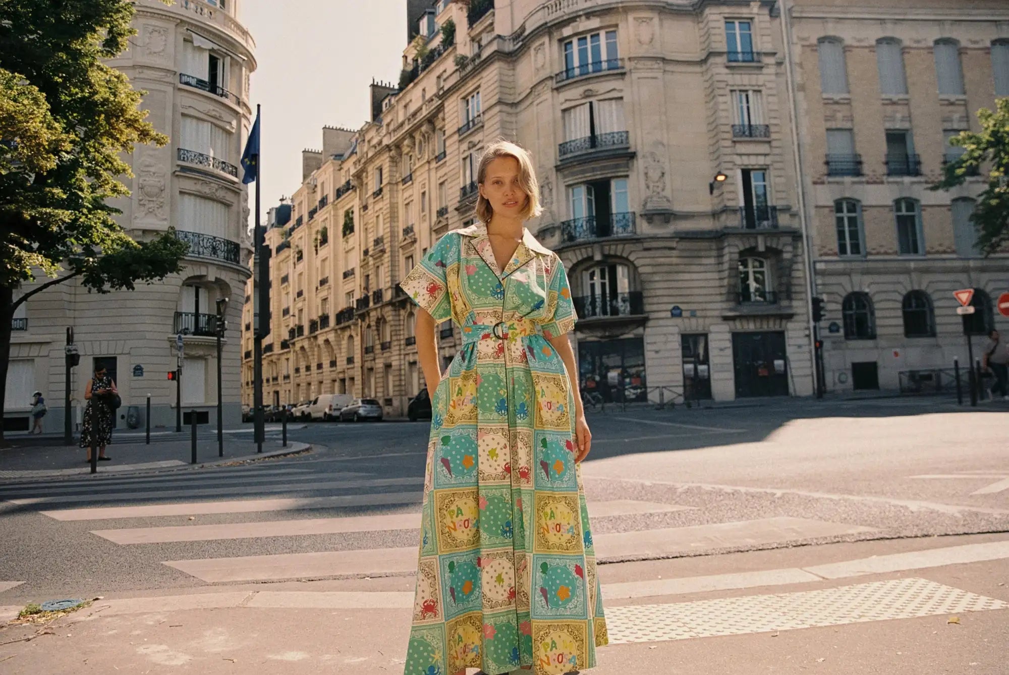 A woman wears a long, vibrant patchwork dress with colorful geometric patterns in shades of green, yellow, and blue.